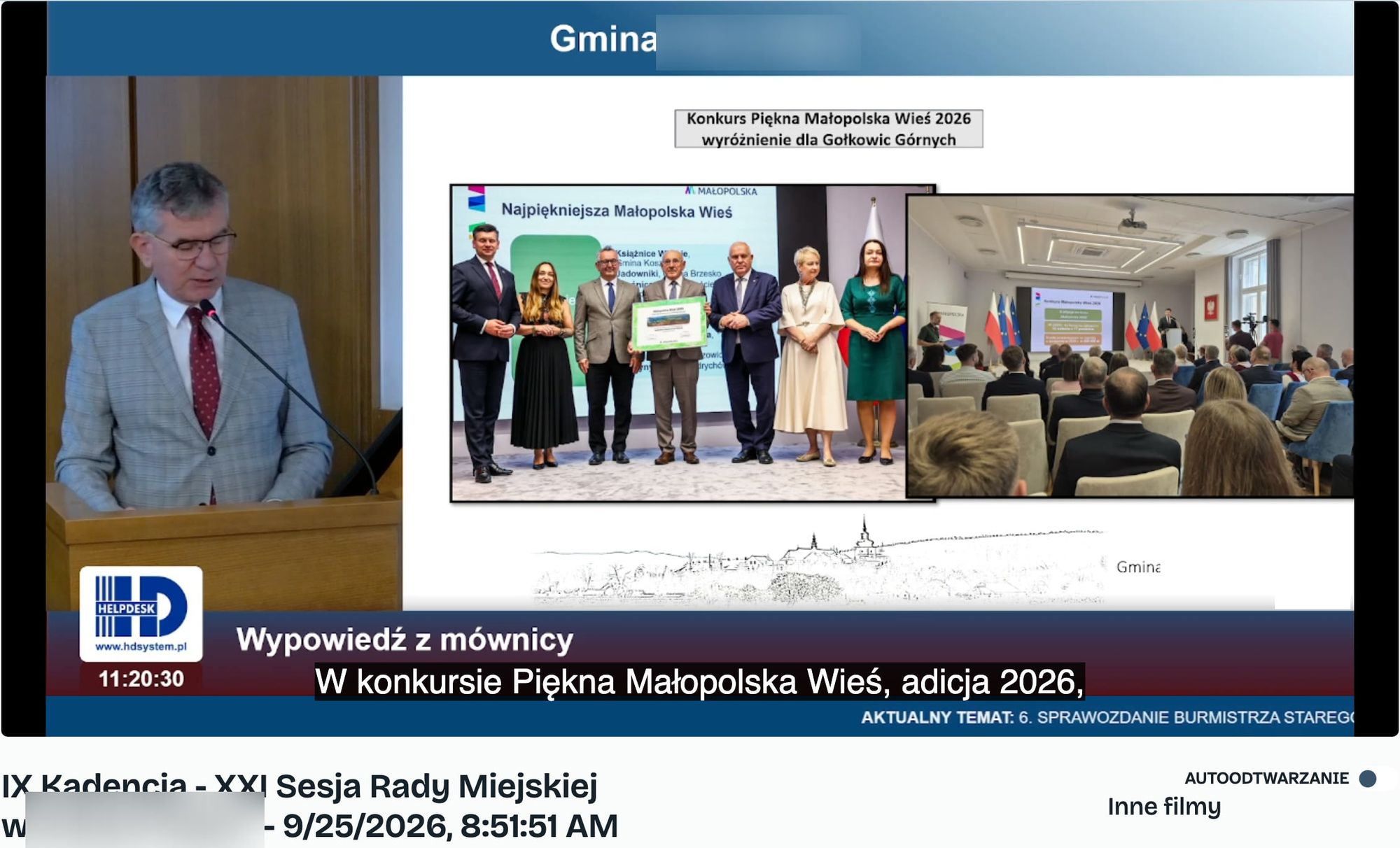 Recording of a council session with automatic subtitles. The mayor at the lectern next to a slide about a regional village contest, with a recognition error in the subtitle at the bottom